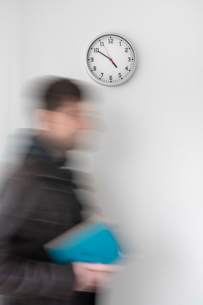 A clock is mounted on the wall, while a person walks in front of the clock. The person is out of focus and appears to be disappearing.