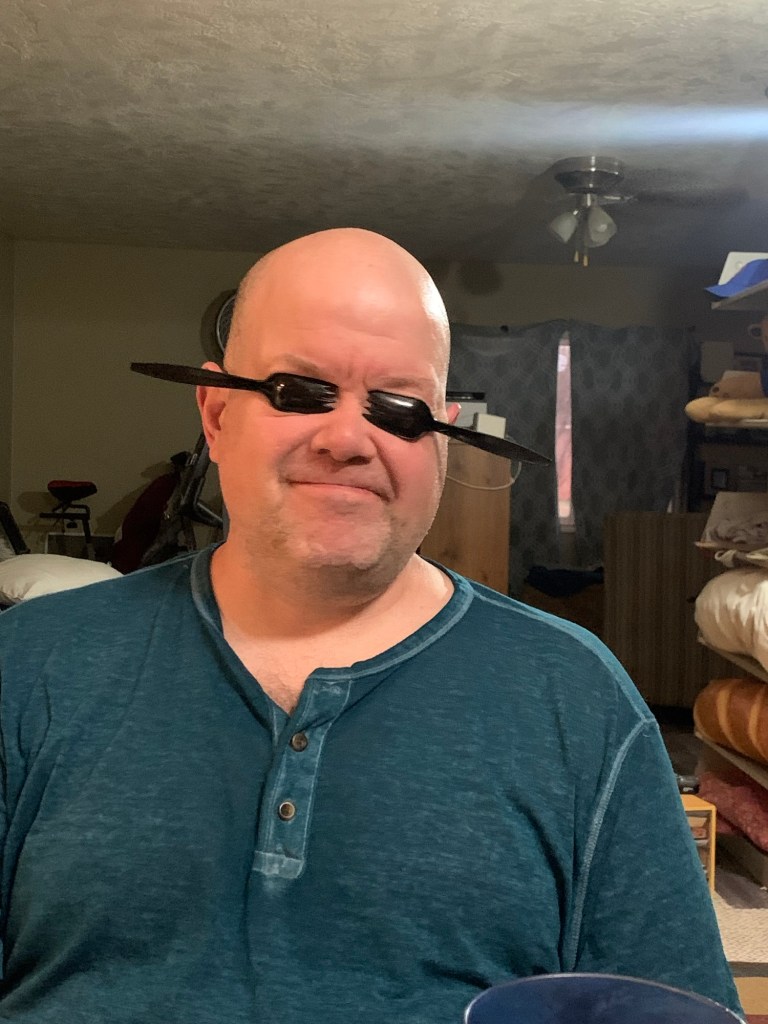 Aaron has a spork covering each eye socket. He is also scrunching his face, to secure the sporks, so that he doesn’t have to use his hands to hold each of them.